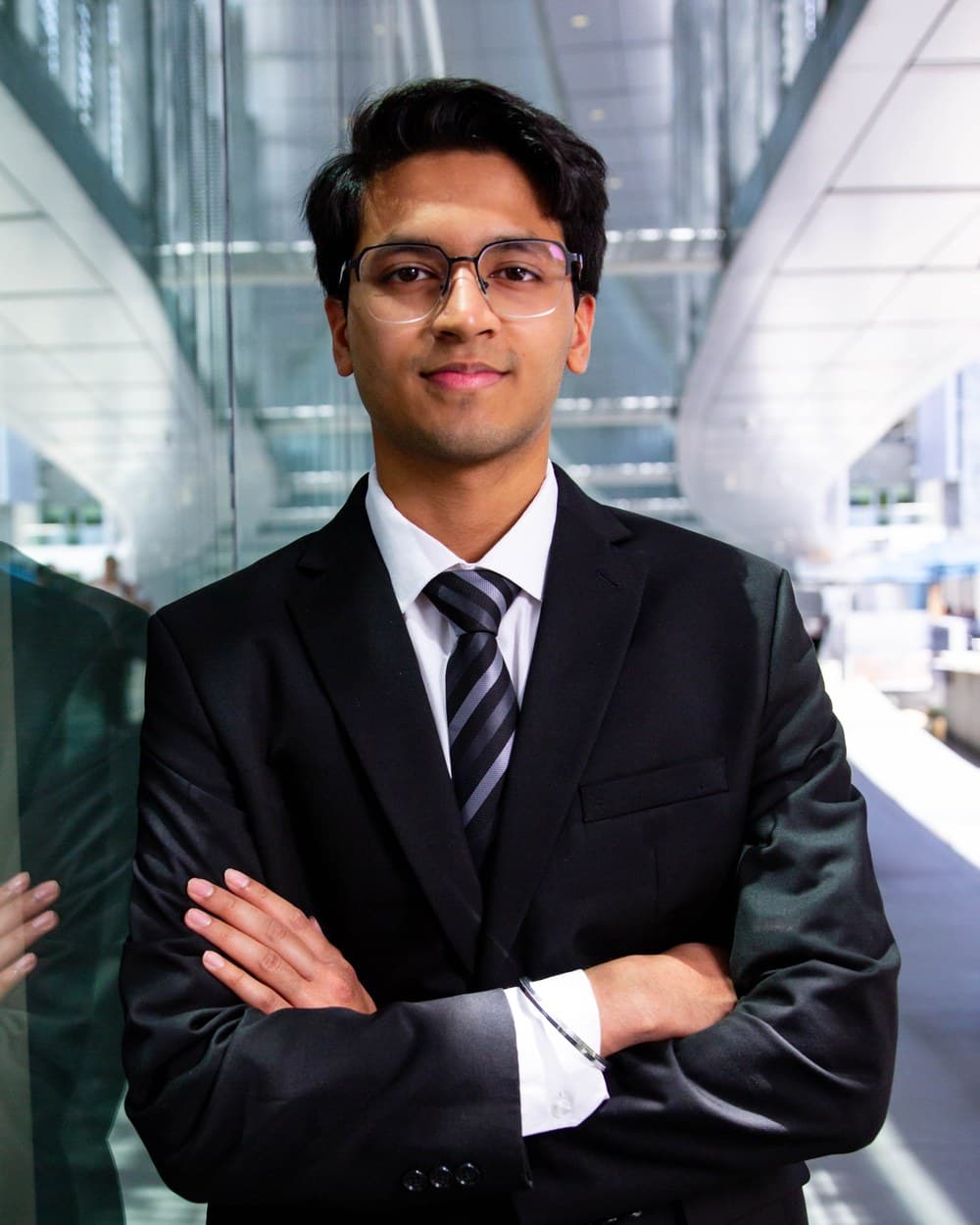Divyam Gupta in a dark suit and striped tie, arms crossed, in a glass-walled atrium